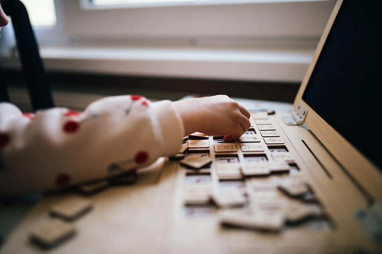 A young child plays with wooden letters on a desk, focusing intently on arranging the pieces. Sunlight streams in through a nearby window, creating a warm atmosphere.